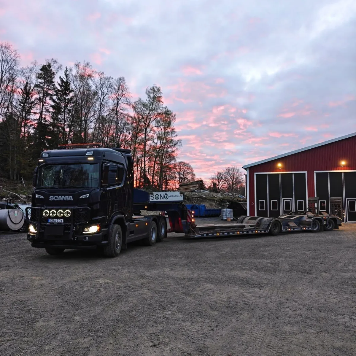 Svart Scania med lågbäddstrailer framför en röd maskinhall i solnedgång